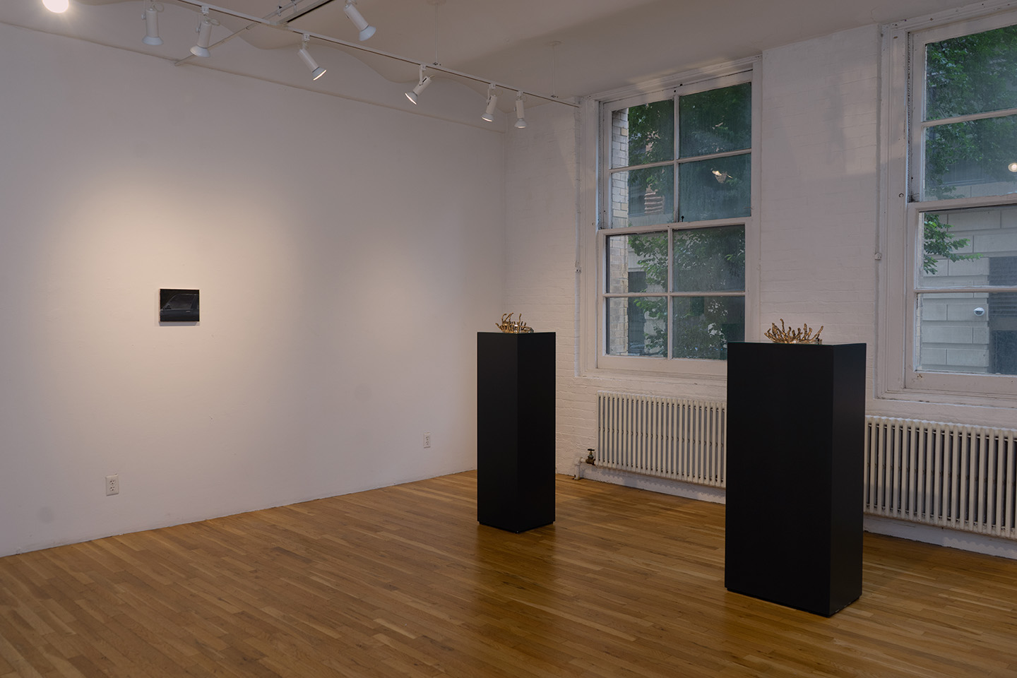 [A view of three artworks in a gallery with two windows. On the left is a small dark painting alone on a white wall and in the gallery’s center, in front of two windows, are two golden-color sculptures, each sitting on a tall black plinth.]